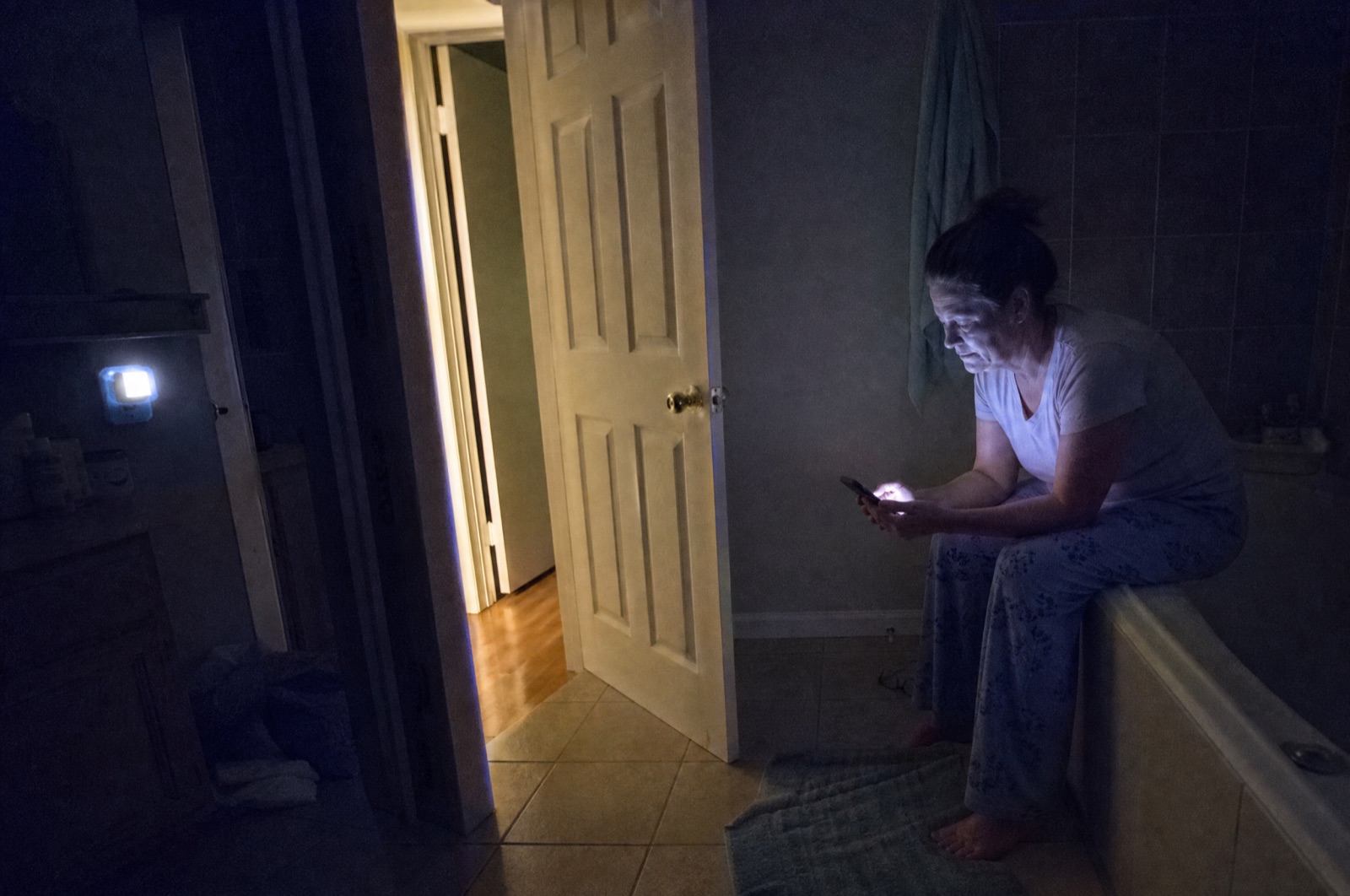 A woman sitting on the edge of the bathtub late at night looking at her phone in a dark bathroom