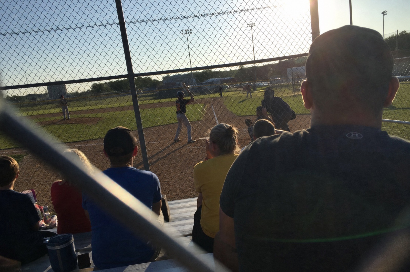 A phone photo taken from the bleachers at a youth baseball game in late afternoon sun